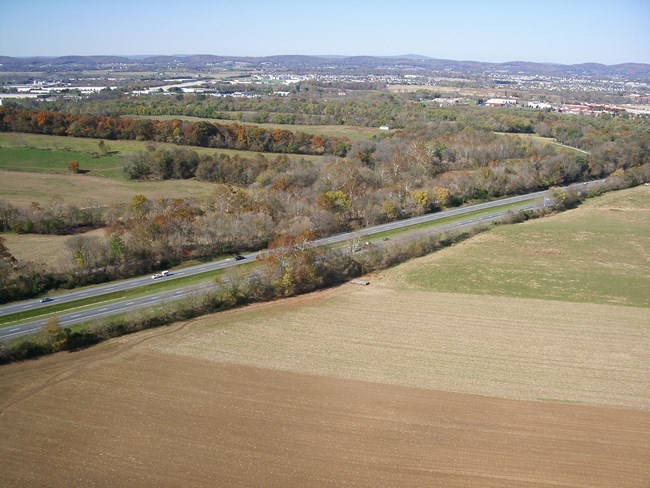 A divide 4-lane highway with farm fields on both sides and buildings in the distance.