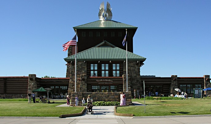 A large stone and wooden building stands above a green grassy lawn with numerous people walking around and a woman with a stroller.