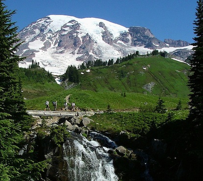 Hikers crossing a bridge over Myrtle Falls in front of Mount Rainier. 