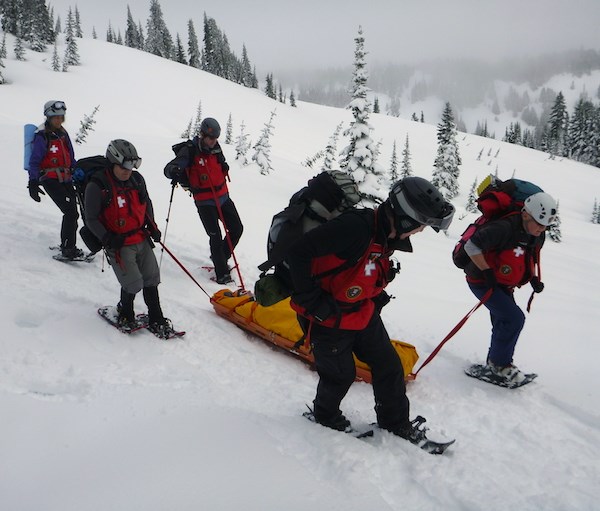 Five people in matching red winter jackets and on snowshoes pull a person wrapped in a yellow blanket on a toboggan across a snowy slope.
