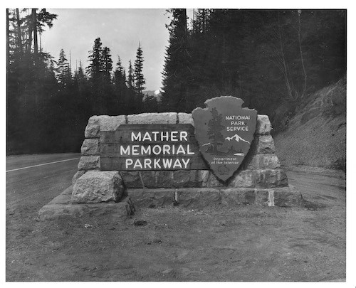 Black and white historic image of a wood sign reading "Mather Memorial Parkway" and a National Park Service Arrowhead attached to a stone base next to a road.