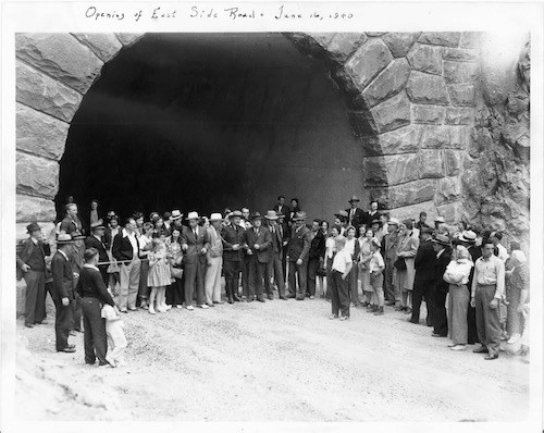 Black and white historic photo of a large group of people, mostly wearing suits or dresses, standing around the entrance to a large tunnel with a stone-clad portal. A ribbon stretches across the entrance to the tunnel with a park ranger holding a pair of