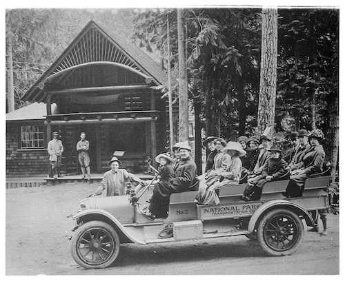 Black and white historic photo of an open-topped vehicle with four row filled with people in early 1900s clothing parked in front of a small log building with a covered porch.