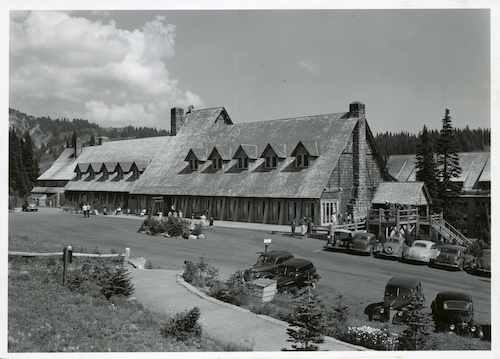 Black and white historic photo of a large wood building with a shingle roof, dormer windows, and stone fireplaces. Old cars line the road in front of the building.
