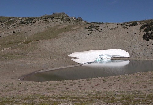 A small lake with a small bank of snow on one edge in a dry rocky valley.