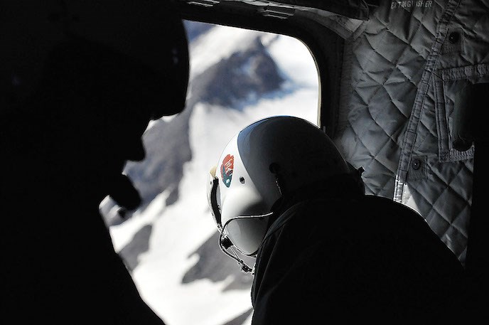 A person wearing a flight helmet with a NPS arrowhead on it leans out the open door of a helicopter.