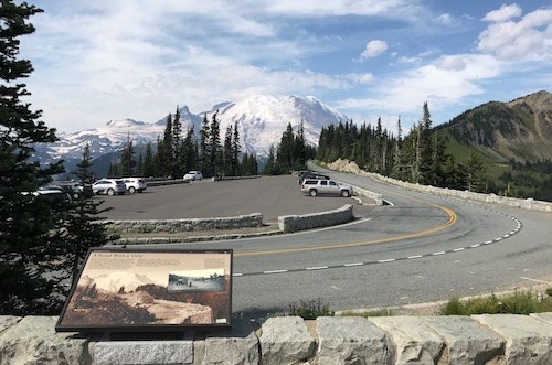 A wayside panel sits on a low rock wall that wraps borders a curved road that wraps around a small parking area with a few vehicles. The road heads away down a ridge towards a glaciated mountain peak.