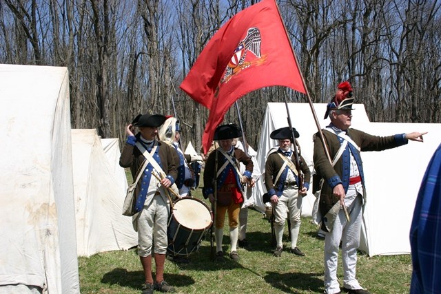 Men dressed in a variety of Continental Army uniforms amid white tents pitched in a field, holding equipment including a drum and a regimental flag.