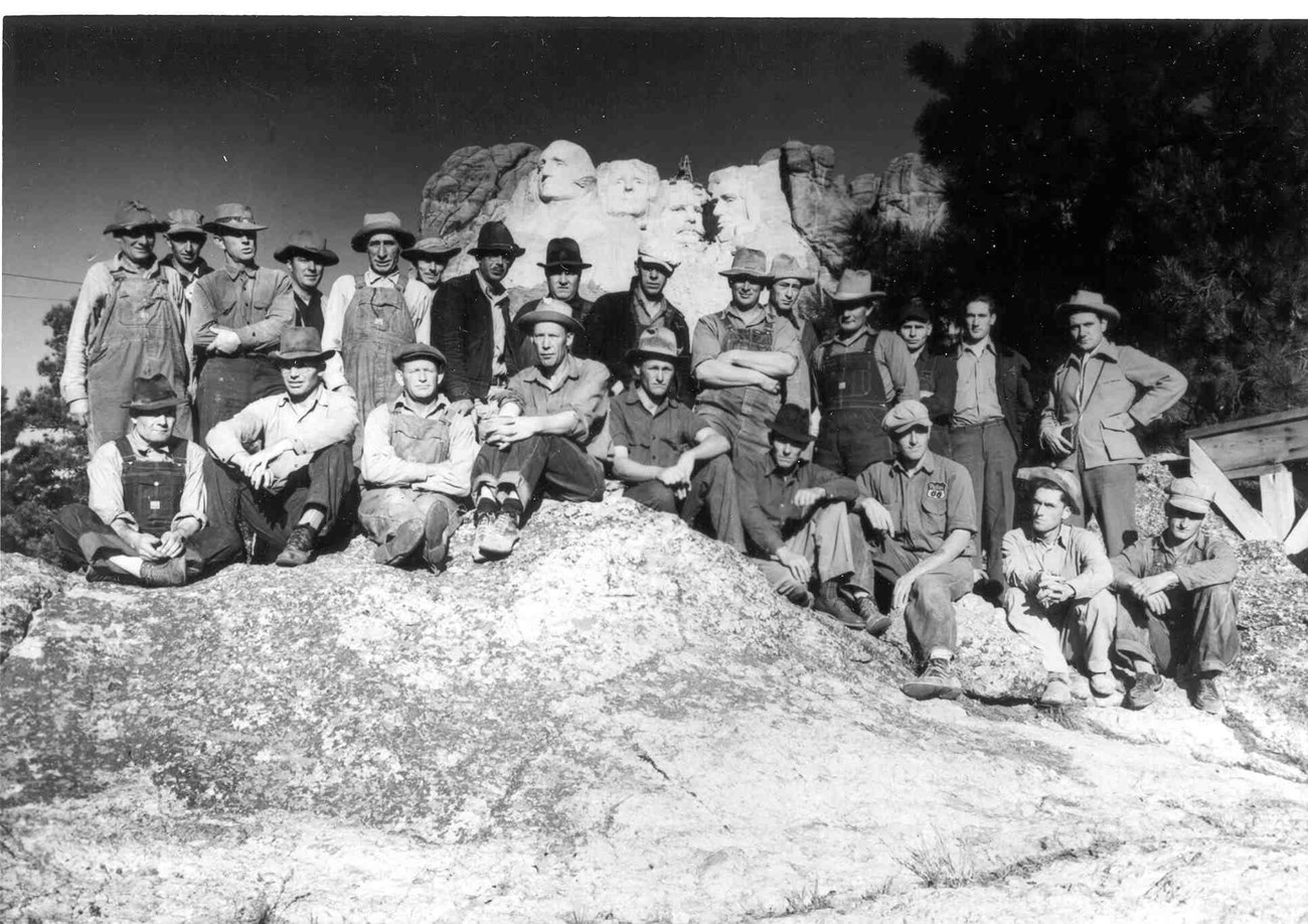 Black and white image of 24 workers in two rows, one seated, one standing on a large rock with Mount Rushmore in the background.  They are all white males wearing hats and clothing suitable for work - overalls, long sleeve shirts and boots.