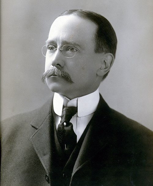 Black and white image of Charles E. Rushmore, a white male with dark hair and mustache.  He wears glasses, a dark suit and tie with a white shirt with a high collar.  He is looking to the left of the image.