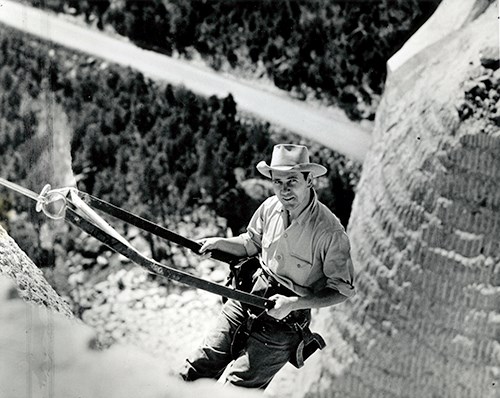 Lincoln Borglum, looking toward the photographer with a partial smile in a bosun chair on the side of Mount Rushmore.  Lincoln is a white male wearing dark work pants, a light-colored button up shirt with the sleeves rolled up and a light-colored fedora.