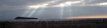 Crepuscular sun rays illumminate a dark island and gray water just above the irregular wooden stockade enclosing the depot at Grand Portage.