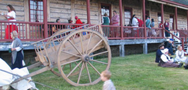 An oxcart rests in short grass while colorfully dressed reinactors enjoy Rendezvous Days on the front porch of the Great Hall.