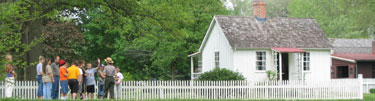 A park ranger with a class of elementary school students outside a white cottage with a white picket fence.