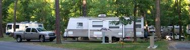 Large white rv from side with silver truck to its left; several other rvs in background. Trees shading campsites.