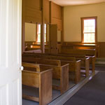 Rows of wooden benches in a sunlight room are seen through a door.