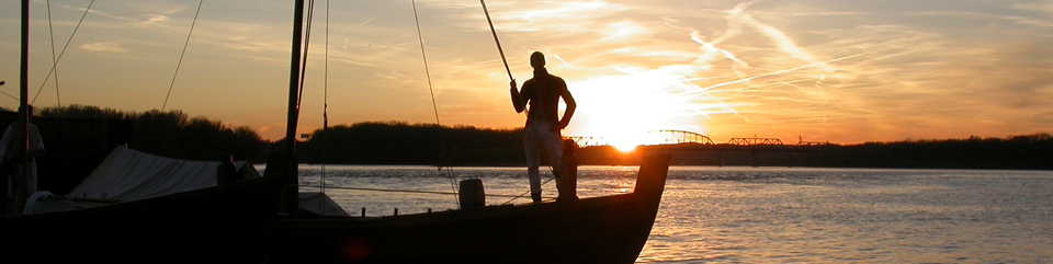 photo of a replica keelboat with a crew member on the bow at sunset