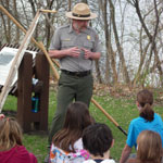 Ranger in uniform stands facing children with logging tools behind him