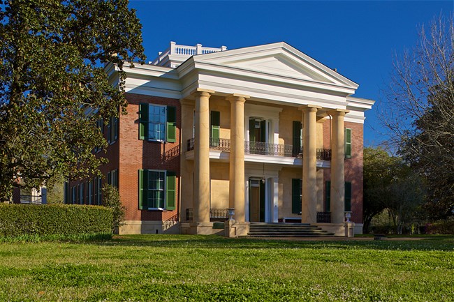 brick mansion with four large columns set resting in informal landscape with lawn and magnolia trees.
