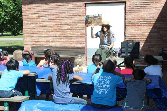 A park ranger sitting on a picnic table in front of a brick wall outside reading and showing a book to young students sitting on picnic tables with blue tarps over them.
