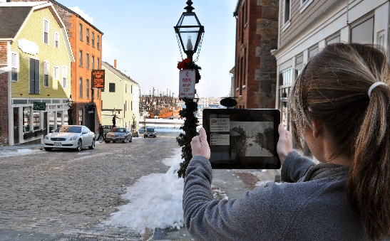 Girl holding an ipad looks at a historic image of the same street she is standing on now.