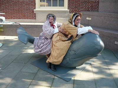 Ruth & Abby in 1850s dress on a whale sculpture smile in jest as the ride the whale.