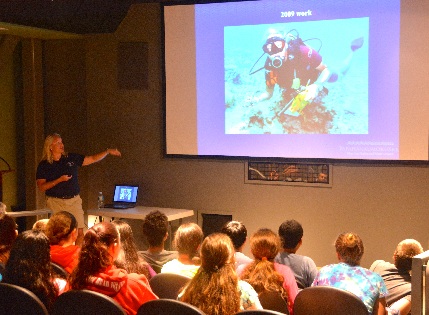 Dr. Kelly Gleason, maritime archeaologist from NOAA and New B Under the Sea project partner, speaks to 33 Sea Lab students during Ocean Day in the national park theater. An image of her writing on her underwater clipboard in on the screen.