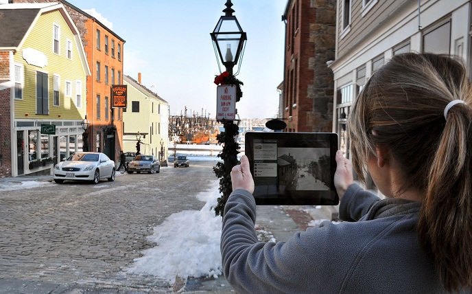 A young lady holds up an ipad with a historic view of Centre Street and actively compares it with the present day view in front of her.