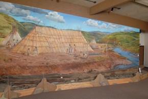 A museum display of a canoe with a mural of a Nez Perce fishing village with a large hut and tipis along a river as a backdrop.