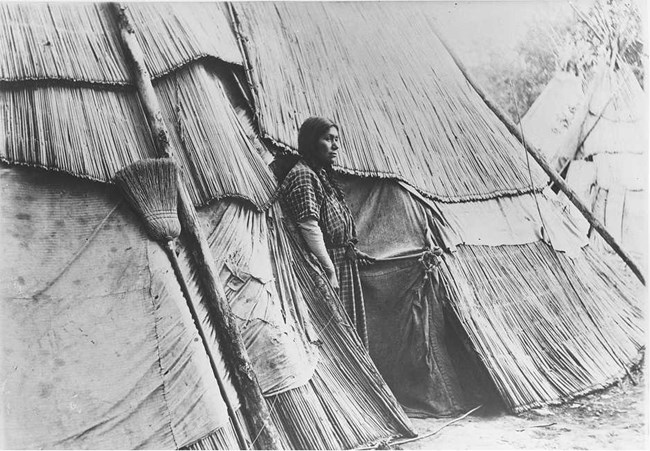 Woman stands in front of a tule lodge.