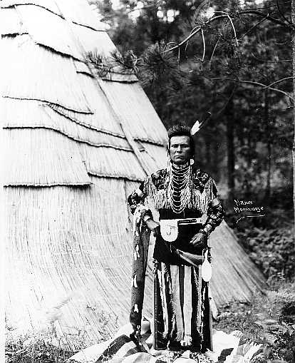 Man in front of tule mat structure dressed in traditional Nez Perce clothing.