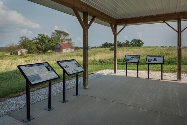 Four interpretive panels mounted under a covered, open-sided kiosk overlooking a prairie landscape containing a few small cabin-like structures.