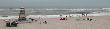 Beachgoers, most in jackets or wrapped in their towels, watch high surf on a lifeguarded beach.