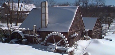 Weathered mill buildings and waterwheels are covered in snow.