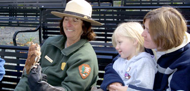 A park ranger, during a Birds of Prey program, holds an owl in her hand as two visitors watch.