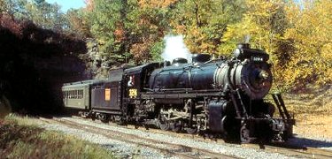 A steam locomotive exits a tunnel pulling olive green passenger cars on one of Steamtown's excursions.