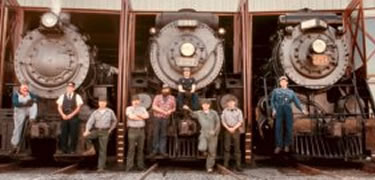 3 steam engines stand in the doors of the roundhouse with crew and mechanics in period and modern dress posing with them.