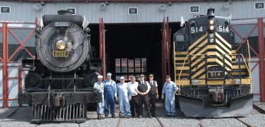Conductors and engineers, in historic dress, pose between a steam engine and a diesel locomotive outside the doors of the Roundhouse.