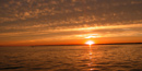 Fiery orange-red sun sets over rippled water, which relects the golden clouds above.