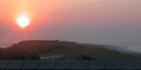 Coral-pink sunrise over low, dark silhouette of the island, a sliver of calm bay water to the left and gentle ocean waves to the right.