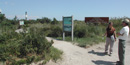 Two people head down sandy trail past bulletin boards and information signs, lighthouse in distance.