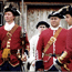 3 soldiers stand at perfect attention. They wear scarlet uniforms, lined with golden rope, and black cocked hats.