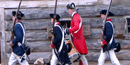 four men march in heavy woolen jackets. one wears a bright red one with a drum at his side, the others blue with muskets in their arms.