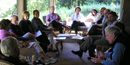 A group of 13 people sit in earnest conversation on the wicker chairs of a sunlit porch, vibrant green woods behind them. NPS Photo.