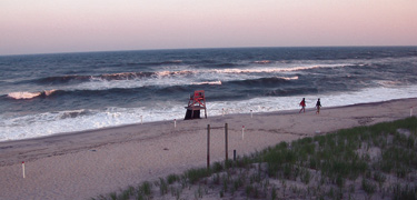 Couple strolls down an empty beach past an unstaffed lifeguard stand, drenched in a pink glow from the sunset to the west.