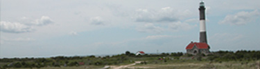 Red-roofed stone house, the keepers' quarters, sits on horizon in front of the white and black banded Fire Island Lighthouse.