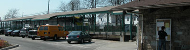 While train pulls into Patchogue railroad station, with cars and orange van parked in front, a waiting passenger reads bulletin board.