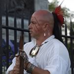 a man in a white shirt, a painted face, and a feathered head-dress stands looking reflectively towards the distance