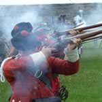 Scarlet red uniformed soldiers are surrounded by lingering smoke as they fire their muskets