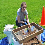 A woman kneels over a screen sifting soil.